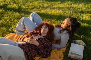 Parkta battaniyede dinlenen iki genç kadın, sıcak günbatımının tadını çıkarırken bira içip kitap okuyorlar, kahkahalar atıyorlar ve birlikte neşeli anılar yaratıyorlar.