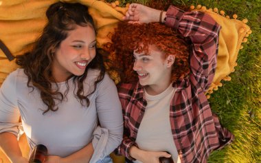 İki genç kadın parkta bir battaniyeye uzanıyor, birlikte rahatlatıcı bir öğleden sonranın tadını çıkarıyorlar, içkilerini paylaşıyorlar ve batan güneşin sıcacık parıltısıyla aydınlanan hoş bir sohbete dalıyorlar.