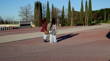 Üniversite parkında birlikte yürüyen, sırt çantalarını taşıyan ve güneş ışığının tadını çıkaran, kaygısız kolej yaşam tarzını paylaşan, arkadaşlığı ve rahatlamış ruh halini somutlaştıran kız öğrenciler.