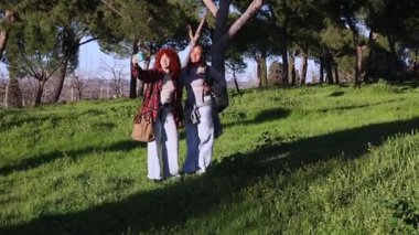 Güneşli park çevresinin tadını çıkarırken selfie çeken kadın gezginler, açık hava maceraları sırasında akıllı telefonlar ve sırt çantalarıyla neşeli anları yakalıyorlar.