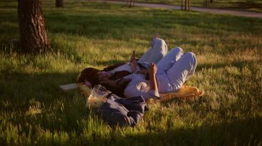 Genç çift parkta battaniyeye uzanıyor, abur cuburları paylaşıyor ve akıllı telefonlar kullanıyor, birlikte bir ağacın gölgesinde rahatlatıcı bir öğleden sonranın tadını çıkarıyorlar.