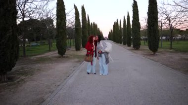 İki genç kadın. Kızıl saçlı ve kırmızı ceketli. Diğeri koyu saçlı ve gri ceketli. Parkta selvi ağaçlarıyla dolu bir yolda yürü ve mutlu bir şekilde konuş. Çanta ve sırt çantası taşıyor.