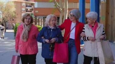 Dört zarif son sınıf kadını, mutlu bir şekilde sohbet ediyorlar ve kol kola geziyorlar, şehir kaldırımlarında geziyorlar, birlikte alışveriş yapıyorlar, renkli alışveriş torbaları taşıyorlar.