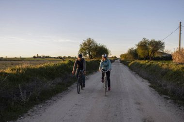 İki bisikletçi sakin bir kır yolunda, ağaçlar ve tarlalarla çevrili, batan güneşin sıcacık parıltısıyla yıkanmış, sakin bir yolculuğun tadını çıkarıyorlar.