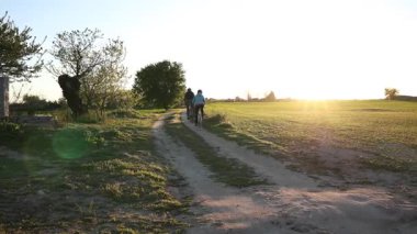 Bisikletçiler altın gün batımında virajlı kırsal yolda pedal çeviriyorlar, uzak ağaçlar ve sıcak akşam ışığı ile çevrili verimli tarım alanları huzurlu bir manzara yaratıyor.