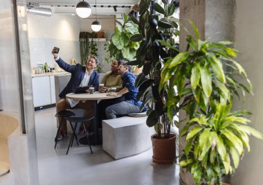 Üç gülen iş adamı modern ofis dinlenme odasında selfie çekiyor, kahvenin tadını çıkarıyor ve yemyeşil bitkilerle çevrili, pozitif bir çalışma ortamı sağlıyor.