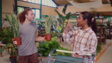 Çok kültürlü ortaklar birlikte ev bitkilerini tarıyor, ev dekorasyonu için yeşillik seçiyor, yerel bahçe merkezinde neşe anlarını paylaşıyor, seçilen bitkileri alışveriş arabasına dikkatlice yerleştiriyor.