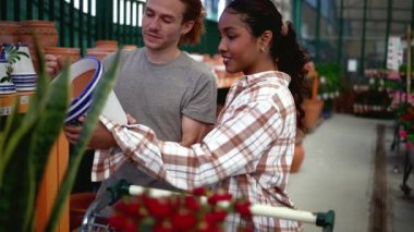 Bahçe merkezinde tarama yapan çok ırklı genç bir çift, farklı boyut ve stilleri dikkatle incelerken, aynı zamanda ev işleri projesi için bitki seçiyor.