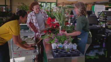 Bahçe merkezi çalışanları müşterilerin bitki ve çiçek seçmesine yardım ediyor, alışveriş ve etkileşim içinde olan farklı bir grup insan, perakende alanında canlı ve canlı bir atmosfer yaratıyor.