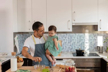 Küçük kızını modern bir mutfakta tutarken önlük takıp bir kaseye yumurta kıran mutlu Latin baba birlikte sağlıklı bir yemek hazırlıyor.