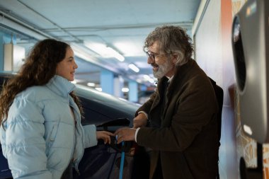 Son sınıf öğrencisi, otoparkta elektrikli arabanın nasıl şarj edileceğini gösteriyor. Genç bir kadınla temiz enerji ve sürdürülebilir hareketlilik çözümleri hakkında bilgi paylaşıyor.