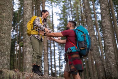 Sırt çantalı yürüyüşçüler bir orman patikasında yol alırken birbirlerine yardımcı oluyorlar, açık havada takım çalışmasının ve desteğin özünü sergiliyorlar. Macera ve bağlantıyı kucaklamak