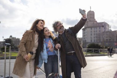 Barselona 'da tatilin tadını çıkaran mutlu bir aile, bavullar ve yeniden kullanılabilir bir kahve fincanı ile selfie çekiyorlar, canlı şehir atmosferinde neşeli anları yakalıyorlar.