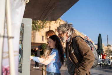 Palma de Mallorca, İspanya 'da güneşli bir günde anne ve büyükbabasıyla interaktif büfeyi işaret eden meraklı kız.