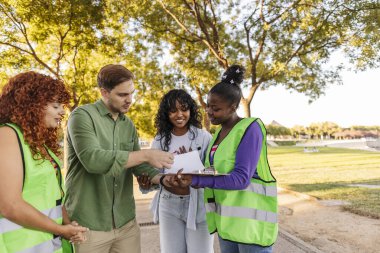Yüksek görüş açısına sahip yelek giyen dört genç gönüllü parkta duruyor, bir pano ve bir kağıt parçasından talimatlar okuyor, bir kamu hizmeti aktivitesine hazırlanıyorlar.