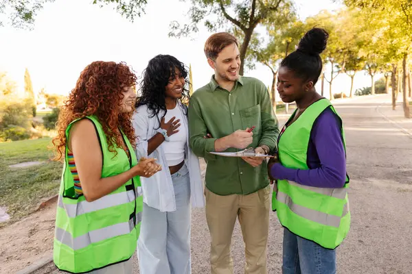 Gönüllüler ve denetçiler parkta bir projeyi tartışıyorlar, güvenlik yelekleri giyiyorlar ve bir pano tutuyorlar, güneşli bir günde takım çalışması ve topluluk ruhunu somutlaştırıyorlar.