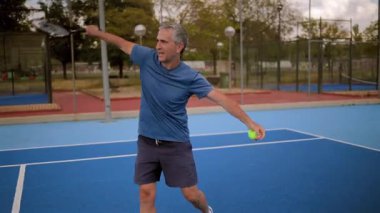 Yaşlı tenisçi, servisini mavi kortta yapıyor. Tekniğinin farklı aşamalarını gösteriyor. Raket ve topu tutmaktan sonraki aşamaya kadar.