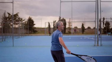 Deneyimli profesyonel tenisçi, parlak mavi kortta güçlü bir servis sunuyor, atletik yetenek ve konsantrasyonu bulutlu akşam gökyüzü altında sergiliyor.