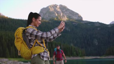 Neşeli genç turistler yaz tatili maceraları sırasında resimli dağ gölü manzarasının tadını çıkarırken selfie anılarını yakalıyorlar.