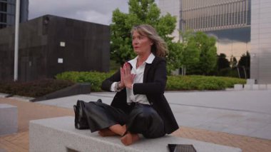 Nilüferde meditasyon yapan kıdemli iş kadını, yoga sırasında tablet kullanarak ve kentsel profesyonel ortamda iç huzuru bularak, kentin iş merkezindeki bankta poz veriyor.