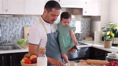 Baba, modern bir mutfakta yemek pişirmek için malzemeler hazırlarken bebeğini kucağında tutuyor ve aile birliği için iç açıcı bir sahne oluşturuyor.