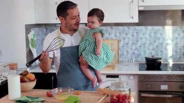 Babası önlük giyip kızını tutarken modern mutfakta cam bir kasede yumurta çırpıyor ve tahta kesme tahtasına meyve ve süt koyuyor.