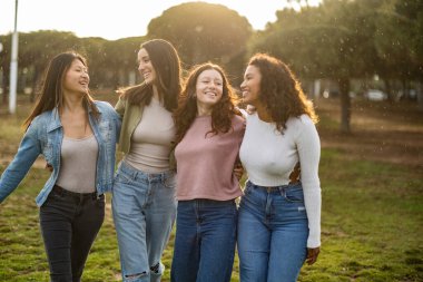 Parkta güneşli bir günün tadını çıkaran dört farklı genç kadın kollarını birbirine dolayarak gülüyor ve arkadaşlıklarını kutluyorlar.