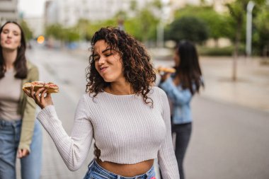 Genç bir kadın arkadaşlarıyla dışarıda pizzanın tadını çıkarıyor. Isırmadan önce lezzetli kokusunu alıyor. Tatmin olmuş bir ifadeyle anın tadını çıkarıyor.