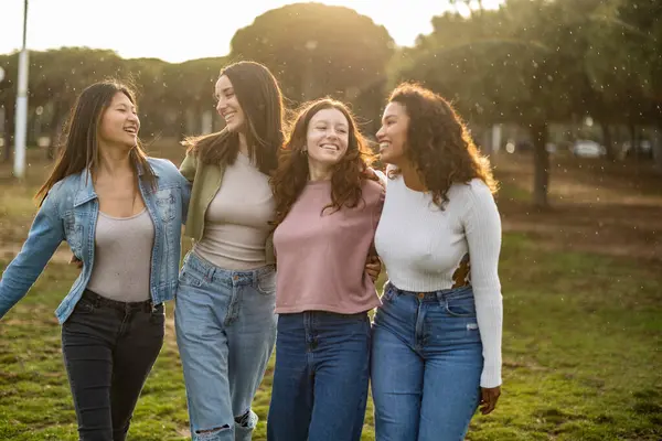 Parkta güneşli bir günün tadını çıkaran dört farklı genç kadın kollarını birbirine dolayarak gülüyor ve arkadaşlıklarını kutluyorlar.