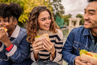 Üç mutlu çoklu etnik arkadaş parkta birlikte oturup lezzetli sandviçlerin tadını çıkarıyor ve kahkahalarını paylaşıyor, boş zamanlarını ve arkadaşlığın güzelliğini kucaklıyorlar.