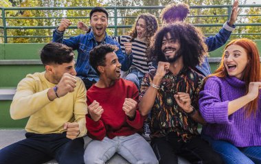 Neşeli çoklu etnik genç öğrenciler parkta tribünlerde oturup, kahkaha ve heyecanı paylaşırken, açık havada arkadaşlığı ve toplumu kucaklarken birlikte kutluyorlar.