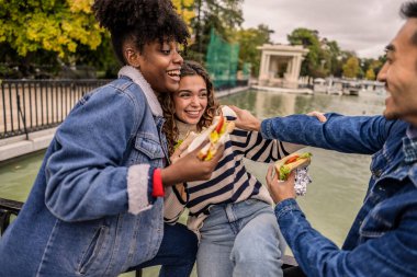 Üç neşeli çoklu etnik arkadaş, birlikte geçirdikleri keyifli tatilde, Madrid, İspanya 'daki sakin bir gölün kenarında dinlenirken gülüyor ve lezzetli sandviçlerin tadını çıkarıyorlar.