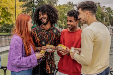 Dört arkadaş nehir kenarında gülüyor ve lezzetli sandviçlerin tadını çıkarıyorlar. Bulutlu bir sonbahar gününde, doğa ve canlı ağaçlarla çevrili neşeli anları paylaşıyorlar.