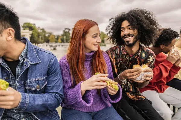 Bir grup neşeli çoklu etnik arkadaş şehir ortamında sandviçleri birlikte yiyor, gülüyor ve hikayelerini paylaşıyor şehrin canlı atmosferini emiyor.