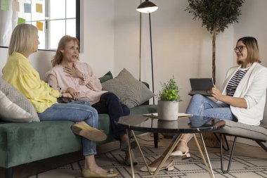 İki endişeli kadın, oturma odasında önemli bir toplantı sırasında elinde dijital tablet tutan bir kadın danışman ile rahat bir koltukta oturup mali konuları tartışıyor.