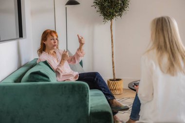 Hasta, modern ofiste bitkilerle ve zarif mobilyalarla terapi seansında el hareketleriyle sorunlarını psikologa anlatıyor.