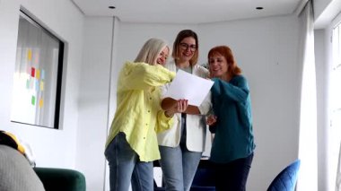 Üç iş kadını birlikte duruyor, bir belgeyi dikkatle inceliyor ve tartışıyor, ortak ofis ortamında düşüncelerini ve görüşlerini paylaşıyorlar.