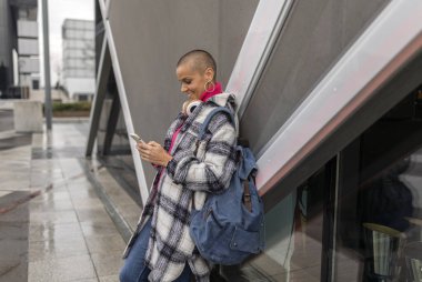 Kulaklık ve sırt çantalı şık bir serbest meslek sahibi, akıllı telefon kullanıyor, müzikten hoşlanıyor ve şehir ortamında modern bir binaya yaslanırken uzaktan çalışıyor.