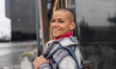 Tıraşlı saçlı, kareli ceket ve pembe boğazlı kazak giyen mutlu iş kadını, şehirde sırt çantası ve kulaklıkla yürürken gülümsüyor.