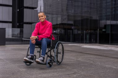 Kafası tıraşlı, rahat kıyafetler giyen, modern ofis binasının dışında tekerlekli sandalyede oturan, gülümseyen ve kameraya bakan kendine güvenen bir iş kadını.