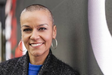 Kafası tıraşlı, kendine güvenen, modern bir ofiste gülümseyen, profesyonellik ve güç saçan, iş yerinde güçlendirme ve çeşitliliği somutlaştıran iş kadını.