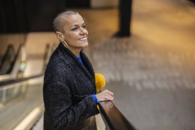 Tıraşlı kafası olan mutlu iş kadını asansörde ya da havaalanında yukarı çıkıyor, palto giyiyor ve elinde sarı çiçekler tutuyor, gülümsüyor ve başka tarafa bakıyor.