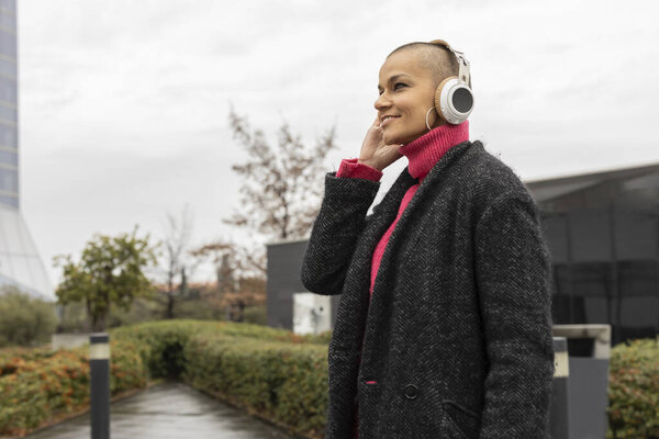 Улыбающаяся деловая женщина в наушниках любит слушать музыку во время прогулки по оживленному деловому району, совершая освежающий перерыв среди современной архитектуры и городской зелени