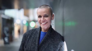 Kendine güvenen, kafası kazınmış iş kadını şehir duvarına yaslanıyor, profesyonel enerji yayıyor ve modern sofistike ve sıcak, güçlü bir gülümsemesi var.