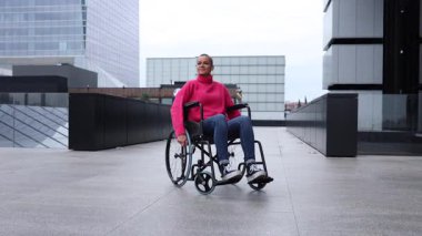 Özürlü genç bir kadın güvenli bir şekilde şehir ortamında geziniyor. Tekerlekli sandalyeyle modern cam binaların yakınına taşınıyor. Güçlendirme ve pozitif yaşam tarzını somutlaştırıyor.