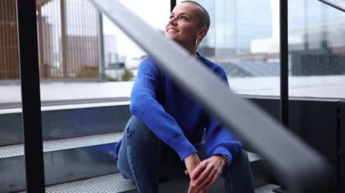 Mavi kazaklı ve kot pantolonlu, kendine güvenen genç kel kadın modern bina merdivenlerinde rahatlıyor, kentli yaşam tarzını gerçek bir gülümseme ve huzurlu bir ifadeyle sergiliyor.