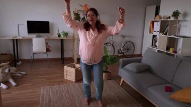 Joyful elderly woman wearing headphones dances and enjoys music in her modern, cozy living room, expressing freedom and happiness in retirement