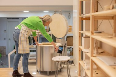 Seramik stüdyosundaki çömlek fırınının sıcaklığını ayarlayan kadın çömlekçi, seramik parçaların sergilendiği raflarla çevrili, el yapımı çömlek yapma sürecini vurguluyor.