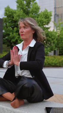 Nilüferde meditasyon yapan kıdemli iş kadını ofis binası yakınında poz veriyor, şehir işi arasında farkındalık uyguluyor, şehir çevresindeki sakinliği ve mesleki sağlığı somutlaştırıyor.