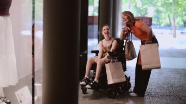 Tekerlekli sandalye kullanan neşeli genç bir kadın ve destekleyici arkadaşı, çeşitli mağazaları keşfederken birlikte alışverişin keyfini çıkarıyorlar, kahkaha ve heyecan paylaşıyorlar.